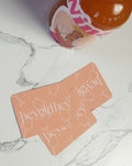 A peach-colored card game with white script text reading 'hey girl hey' and cocktail glass line drawings, placed on a white marble surface next to a partially visible orange bottle.